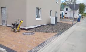 Pomarańczowa kostka brukowa ułożona wokół studzienki kanalizacyjnej przy ścianie budynku, obok ubijarka wibracyjna, widoczne obrzeża z ciemnej kostki i fragment posesji sąsiedniej.