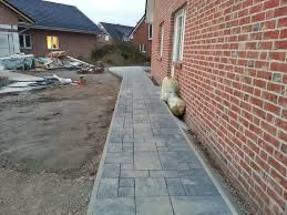 Świeżo ułożony chodnik z szarej kostki brukowej o zróżnicowanych kształtach, biegnący wzdłuż ceglanej ściany budynku. W tle widoczny plac budowy z materiałami i kolejny budynek w trakcie budowy.