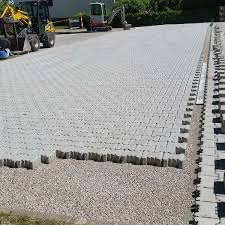 Jasnoszara kostka brukowa ułożona na dużej powierzchni, z widocznymi maszynami budowlanymi w tle i rzędem kostek przygotowanych do ułożenia na krawędzi.