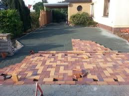 Częściowo ułożona, prostokątna kostka brukowa w odcieniach czerwieni i beżu na podjeździe do domu. Widoczne przygotowane podłoże z czarnej geowłókniny. Fragmenty kostki nieułożone, leżące obok.