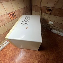 Mirosława Mira - Wnętrze kuchni z brązowymi płytkami i białym piekarnikiem elektrycznym. Widoczne gniazdka i przedłużacz. Zdjęcie zrobione z góry.