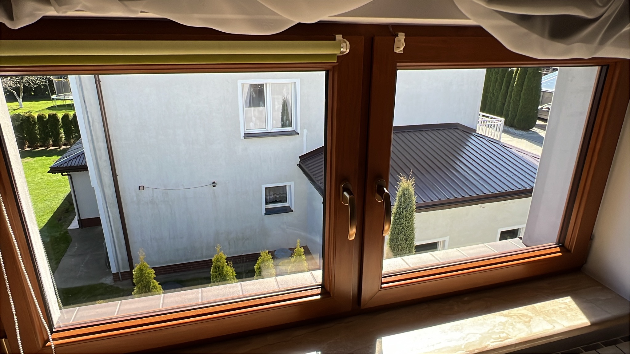 Widok z okna na dom z brązowym dachem, zielony trawnik i ozdobne krzewy, rama okienna z drewna.