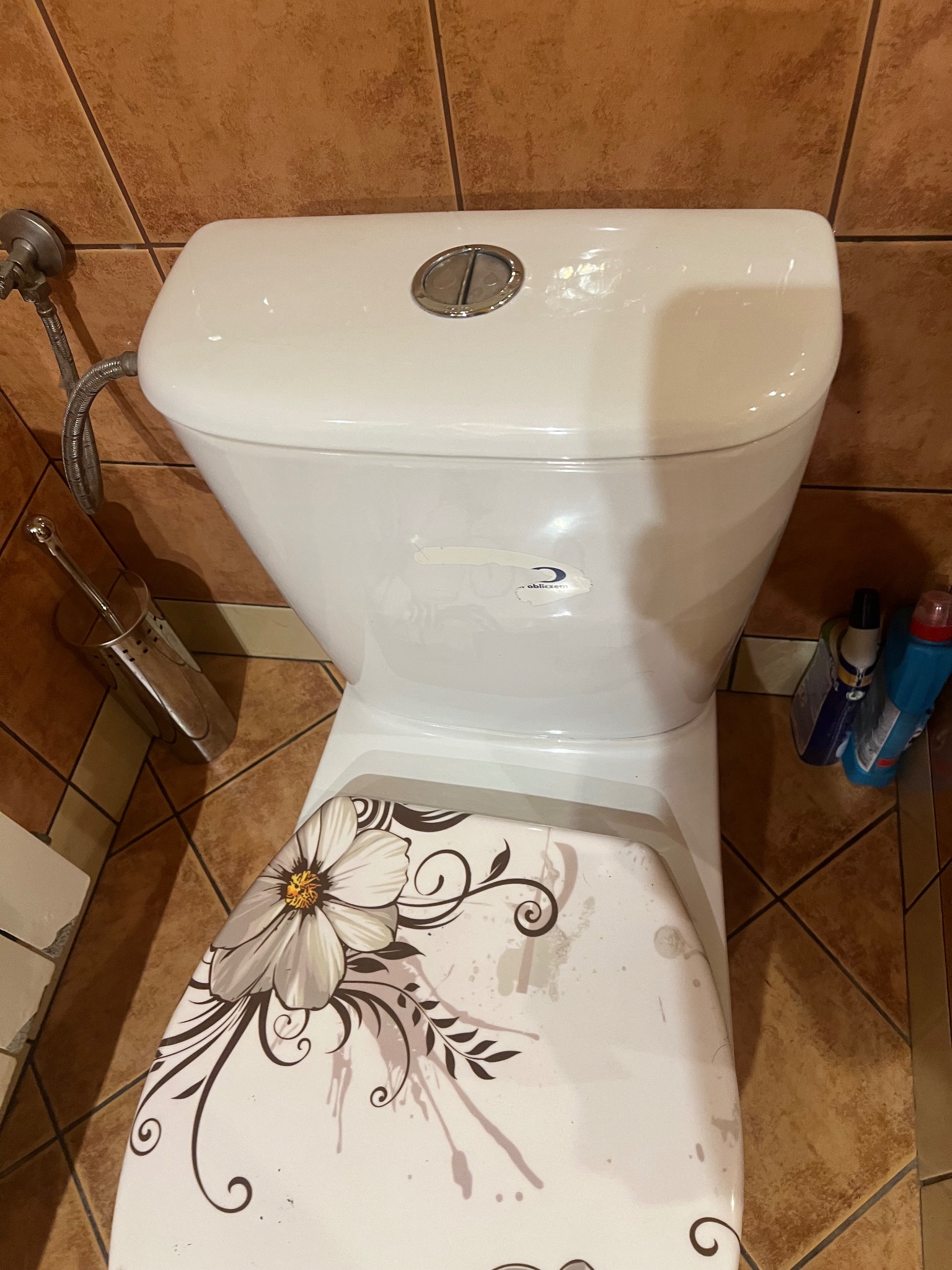 Toaleta z deską ozdobioną motywem kwiatowym na tle brązowych płytek, widoczne środki czystości i szczotka do WC.