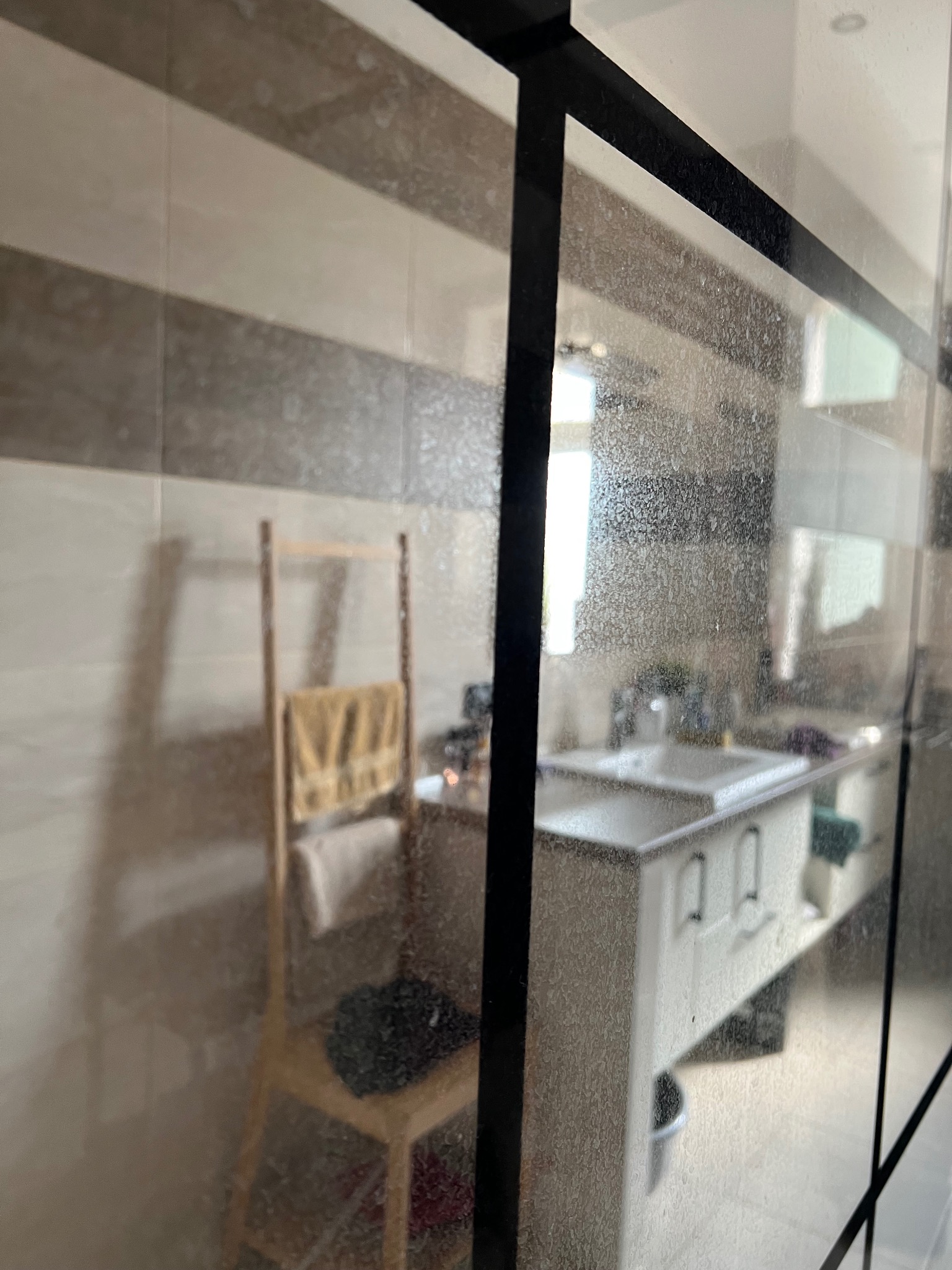 Zabrudzona szyba kabiny prysznicowej z widocznym wnętrzem łazienki: umywalka z szafką, drabinka na ręczniki i fragment ściany z poziomymi pasami płytek.