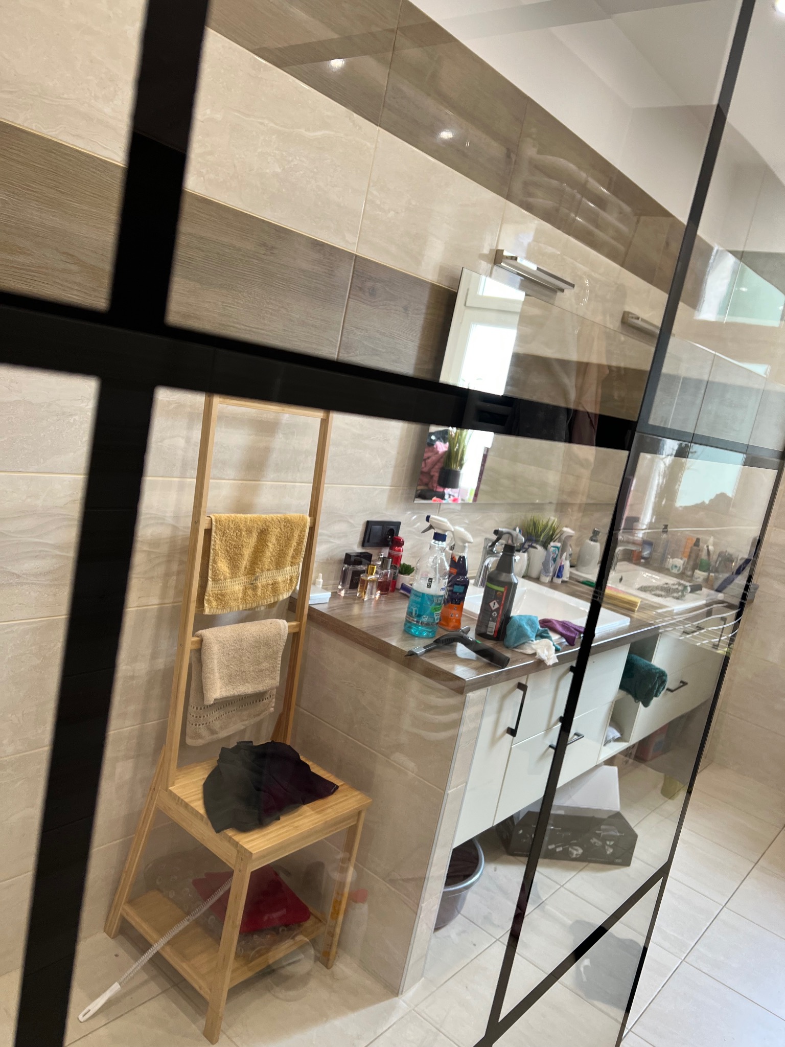 Umywalka w łazience z widocznymi środkami czystości i akcesoriami do sprzątania, takimi jak ściągaczka do wody i ściereczki, oraz drewniany regał z ręcznikami i ubraniami.