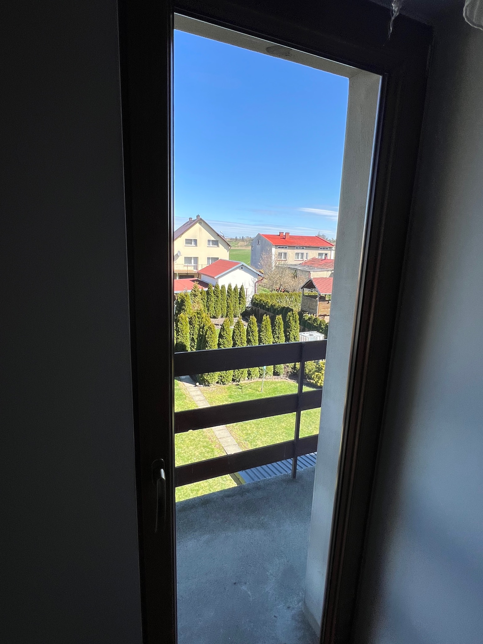 Widok z okna balkonowego na zadbany ogród z równo przyciętymi tujami, trawnik i zabudowania mieszkalne w słoneczny dzień.