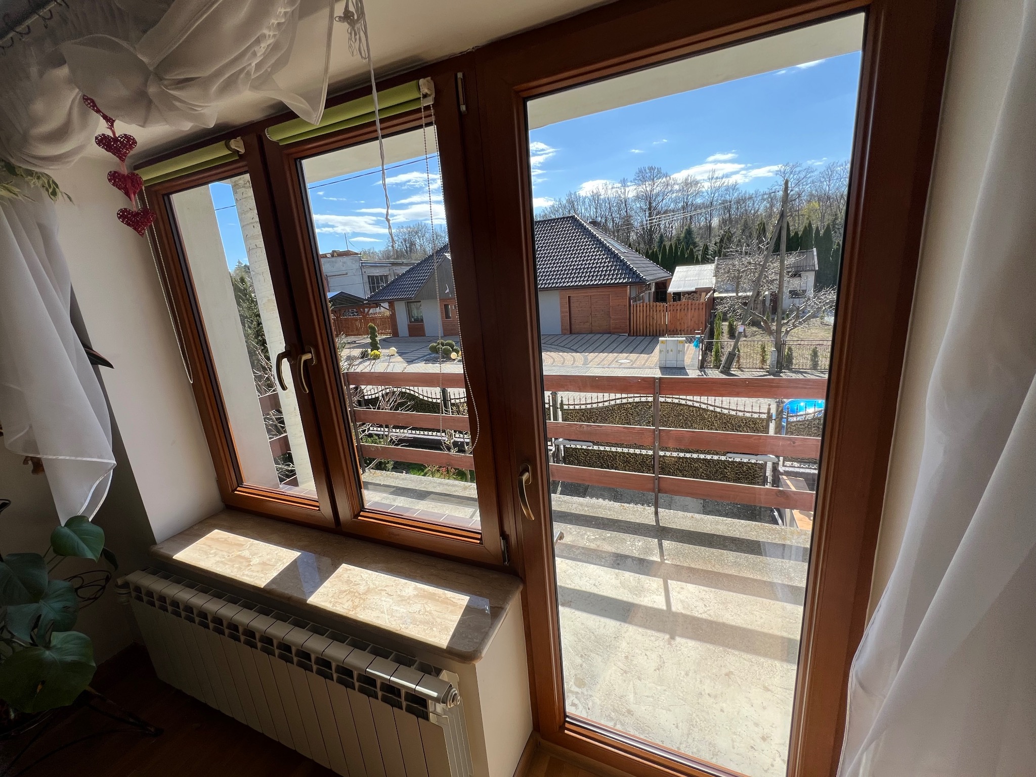 Drewniane okno z widokiem na dom z ogrodem i basenem w słoneczny dzień, firanka z dekoracjami w kształcie serc.