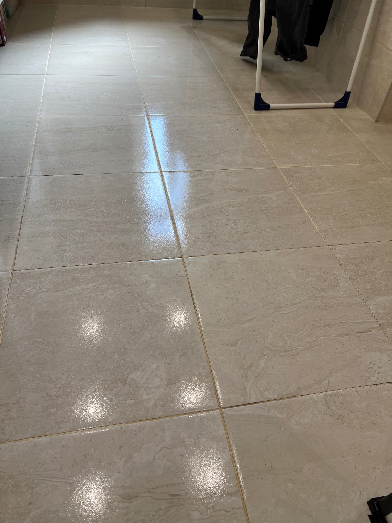 Świeżo umyte, lśniące płytki podłogowe z widocznymi smugami wody i odbiciami światła, fragment suszarki na pranie w tle.