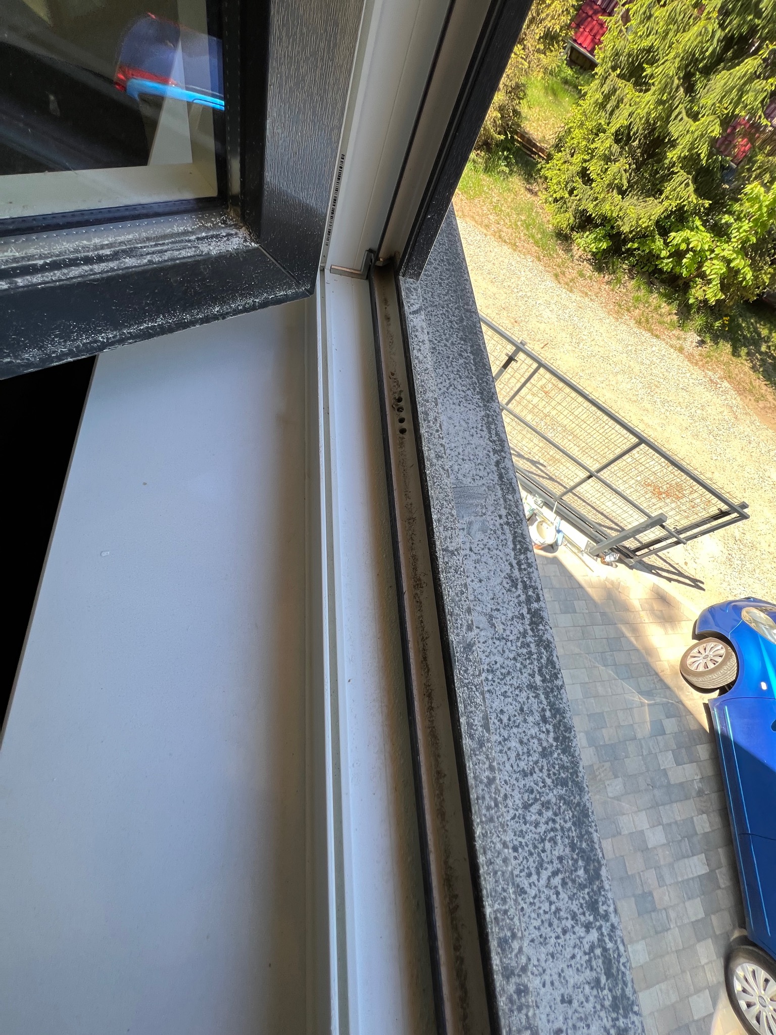 Silnie zabrudzone okno PVC z widocznymi plamami i zaciekami, z zewnątrz widok na podjazd, samochód i zieleń. Konieczność umycia ramy i szyby.