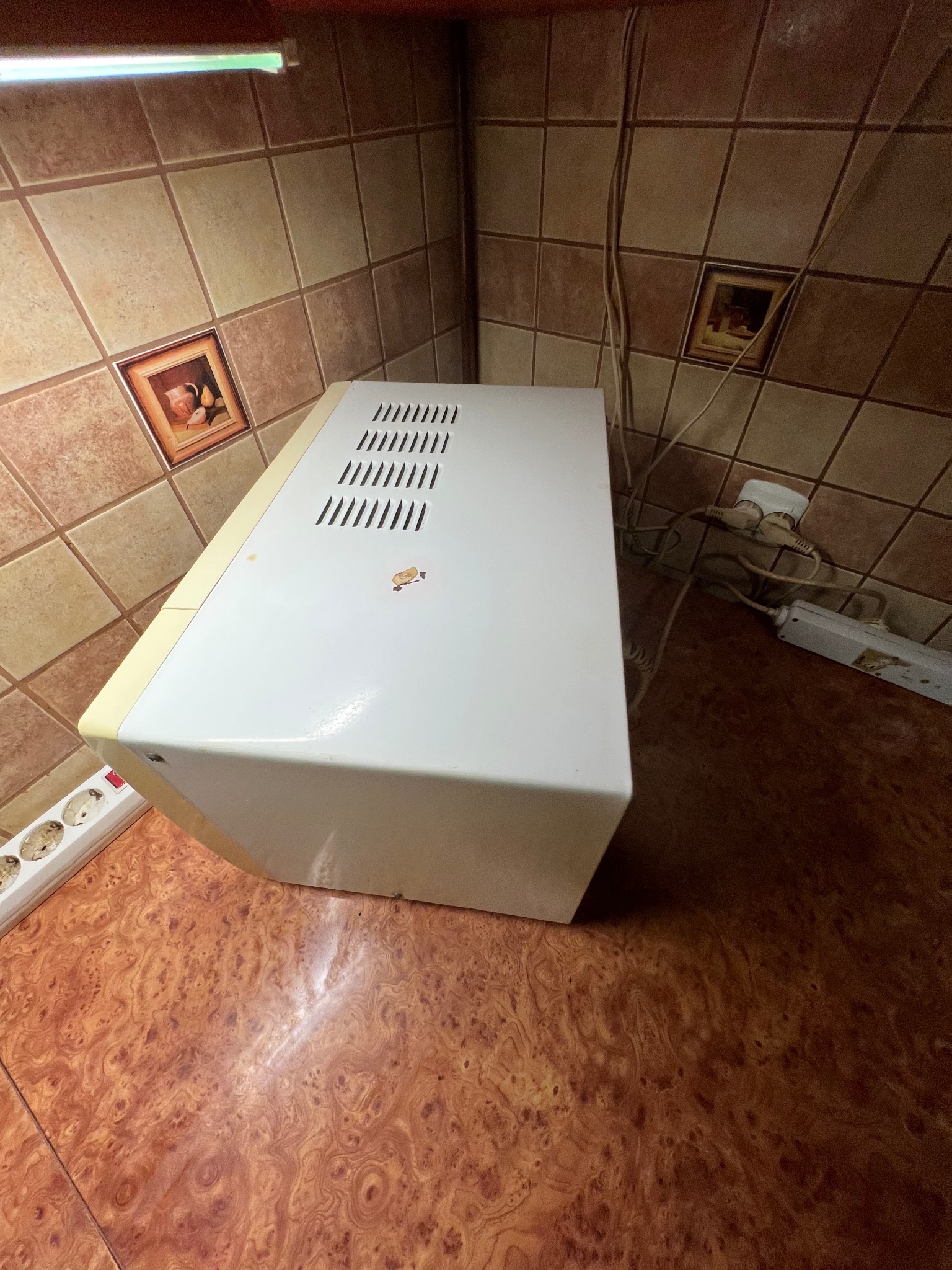 Wnętrze kuchni z brązowymi płytkami i białym piekarnikiem elektrycznym. Widoczne gniazdka i przedłużacz. Zdjęcie zrobione z góry.