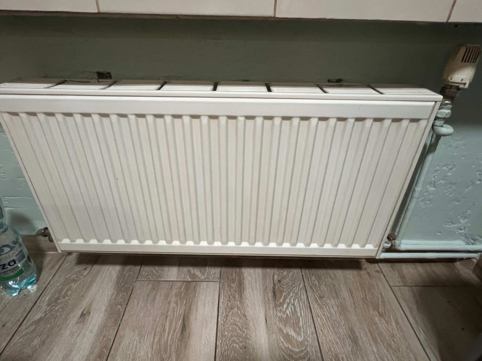 Biały, czysty grzejnik na tle drewnopodobnej podłogi i ściany. Widoczny termostat i butelka wody mineralnej. Zdjęcie po czyszczeniu lub wymianie grzejnika.