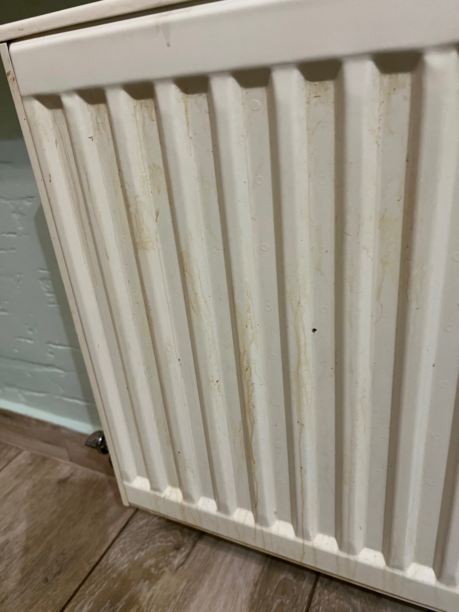 Zabrudzony, biały grzejnik żeberkowy z widocznymi smugami i plamami, stojący na drewnianej podłodze przy jasnej ścianie. Ukazany stan 'przed' czyszczeniem.