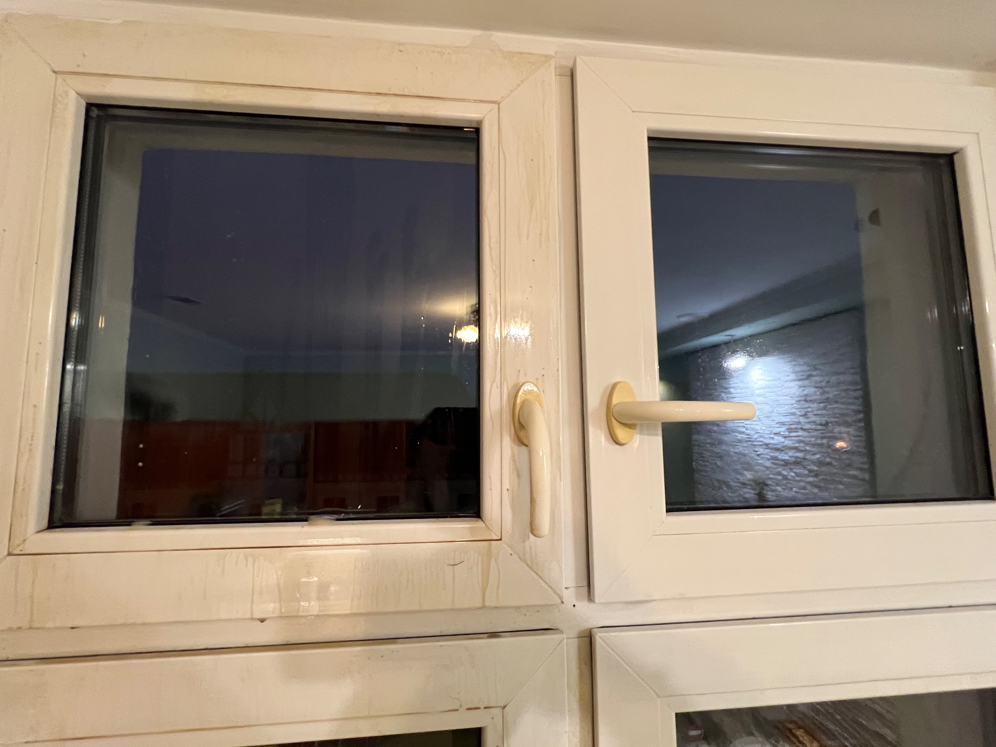 Brudne, białe ramy okienne z widocznymi zaciekami i zabrudzeniami. Okna zamknięte, z widocznymi klamkami. Widok z bliska, ukazujący potrzebę czyszczenia.