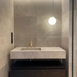 Nowoczesna łazienka z kamienną umywalką, złotą armaturą i kulistą lampą wiszącą. Płytki w odcieniach szarości i podświetlenie LED tworzą elegancki klimat.
