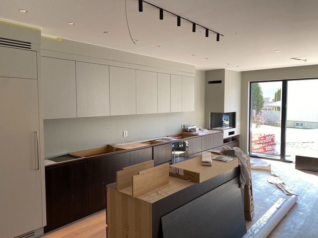 Jasna kuchnia w trakcie montażu: białe szafki wiszące, ciemne dolne, wyspa kuchenna z elementami do złożenia, duże okno z widokiem na zewnątrz. Oświetlenie szynowe.