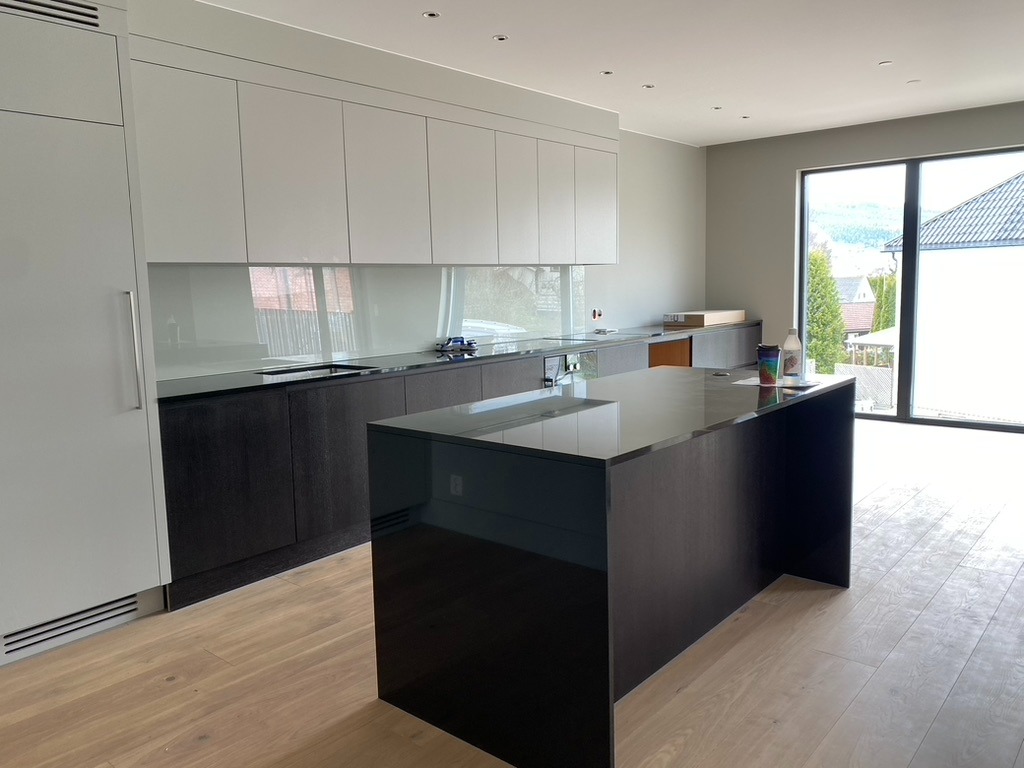 Nowoczesna kuchnia z wyspą: białe szafki górne, ciemne dolne, jasna podłoga. Duże okno z widokiem na zieleń. Minimalistyczny design, czyste linie.