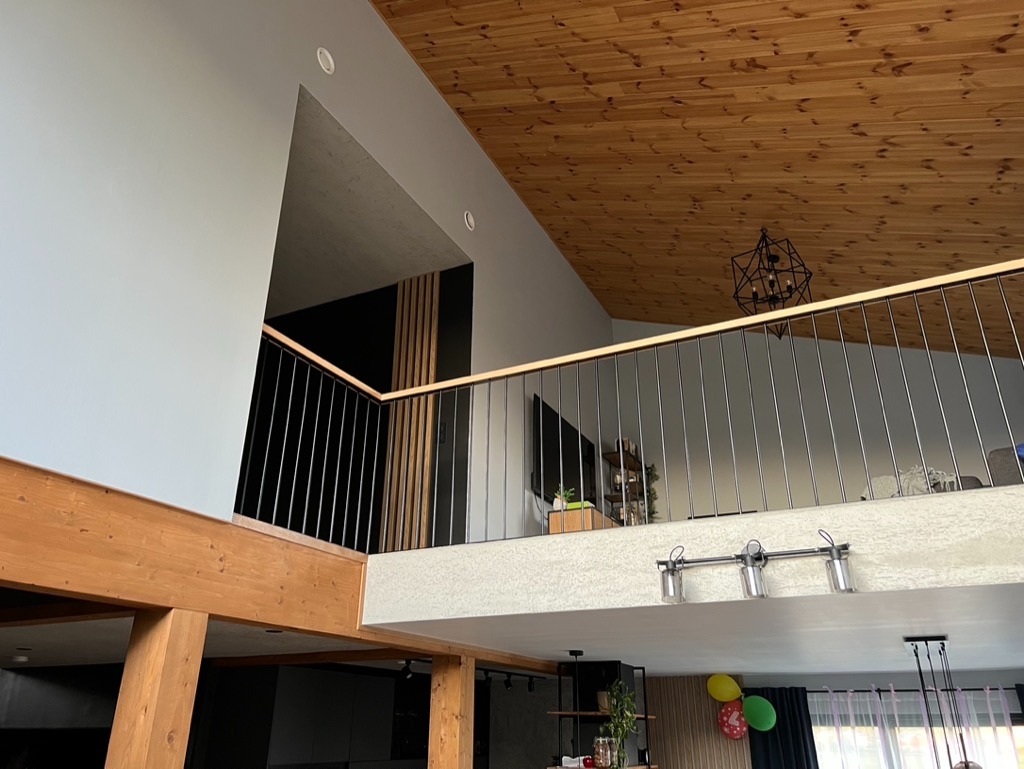 Wnętrze domu z drewnianym sufitem i balustradą. Widoczne drewniane belki, nowoczesne oświetlenie, elementy wykończenia ścian i sufitu. Dwupoziomowa przestrzeń.