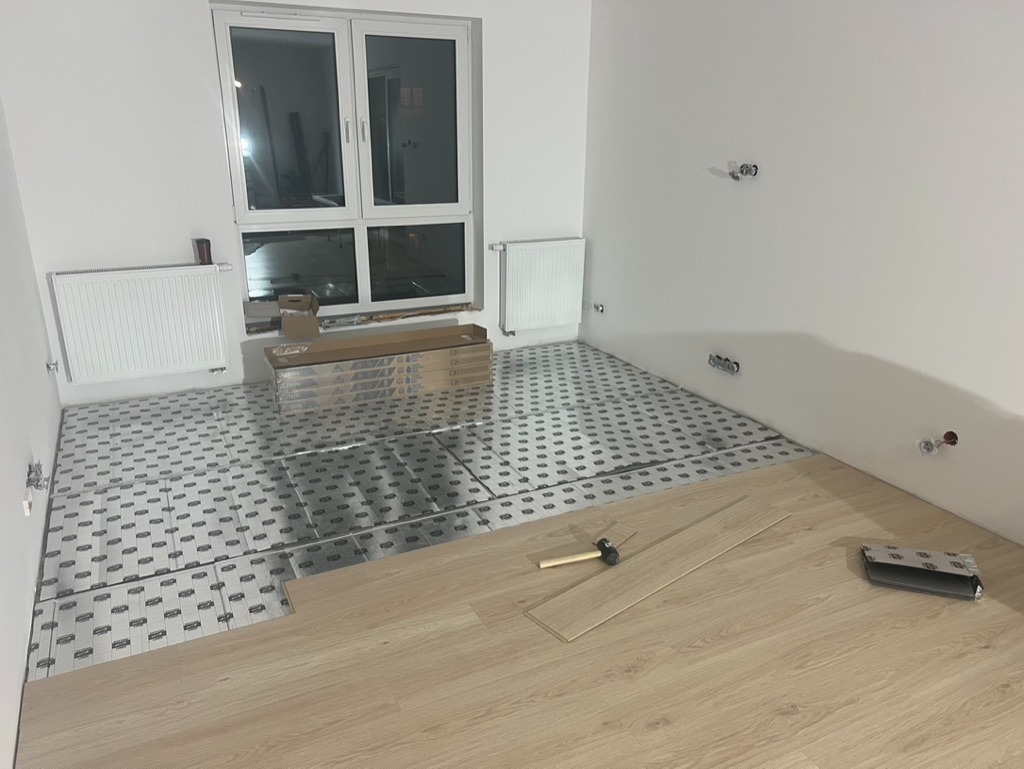 Układanie paneli podłogowych na folii izolacyjnej w jasnym pokoju. Widoczne narzędzia i paczki paneli. Częściowo ułożona podłoga, etap prac wykończeniowych.