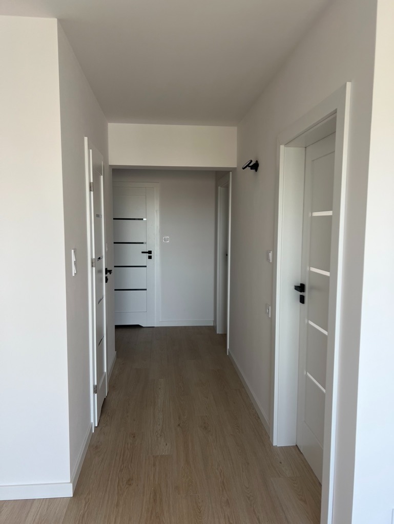 Jasny korytarz z drewnopodobną podłogą i białymi drzwiami z czarnymi klamkami. Minimalistyczne oświetlenie ścienne. Widok w głąb mieszkania.