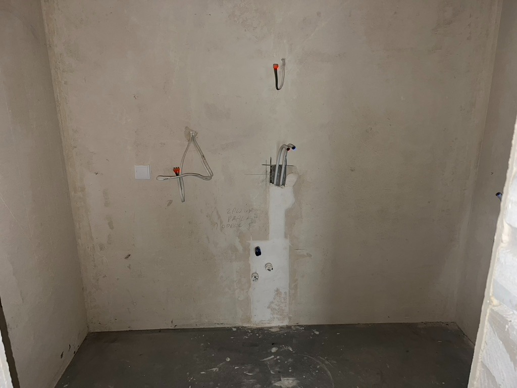 Surowy stan wnętrza: ściany po tynkowaniu z widocznymi instalacjami elektrycznymi i hydraulicznymi przed wykończeniem. Szare podłoże, brak podłogi. Widoczne przewody i puszki.