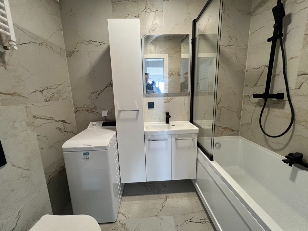 Nowoczesna łazienka w beżowych marmurkowych płytkach z białą armaturą: pralka, szafka z umywalką, lustro i kabina prysznicowa z czarną armaturą.