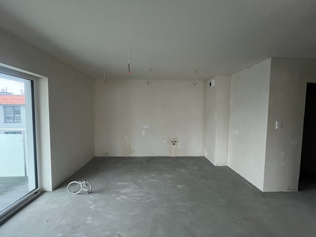 Surowe wnętrze mieszkania po wylewce, widoczne przewody elektryczne i niedoskonałości ścian, przygotowane do dalszych prac wykończeniowych. Okno balkonowe z lewej strony.