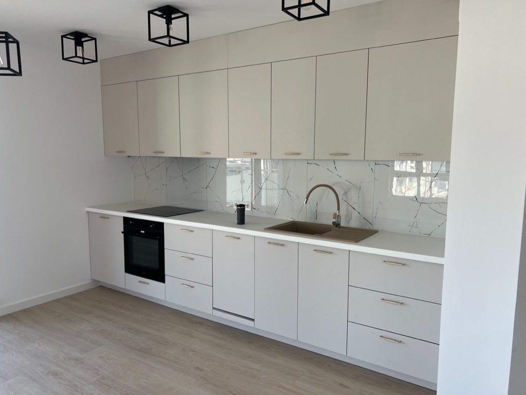 Jasnoszara zabudowa kuchenna z marmurowym backsplashiem i złotymi uchwytami. Nowoczesne oświetlenie sufitowe. Minimalistyczny design.