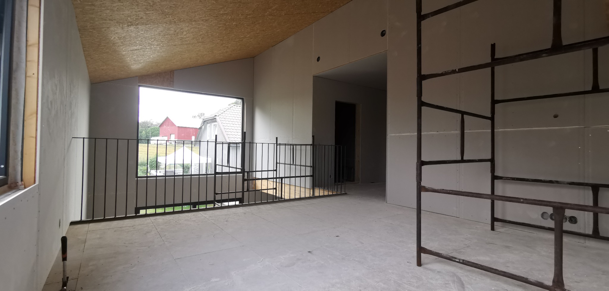 Poddasze w trakcie wykańczania: płyty gipsowe na ścianach, drewniane panele na suficie, metalowa balustrada przy oknie. Widoczne rusztowanie. Surowy stan wnętrza.