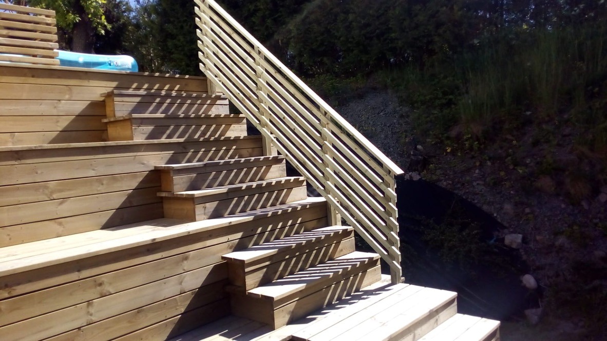 Drewniane schody zewnętrzne z barierką, oświetlone słońcem, prowadzące do ogrodu. Widoczne detale konstrukcyjne i naturalna kolorystyka drewna.