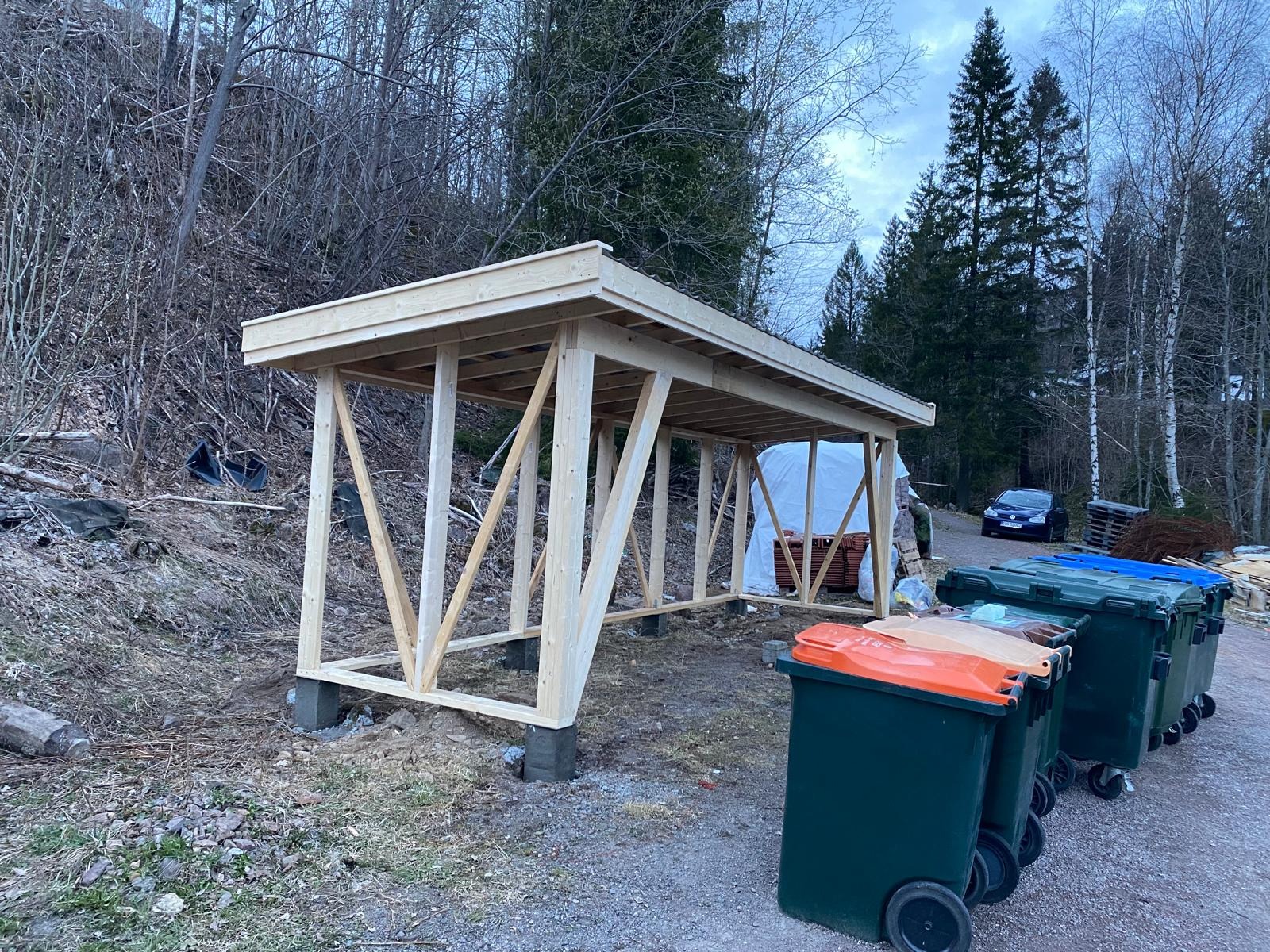 Drewniana konstrukcja w trakcie budowy na zewnątrz, widoczne słupy, belki i zadaszenie. W tle las i pojemniki na śmieci. Surowy stan, brak wykończenia.
