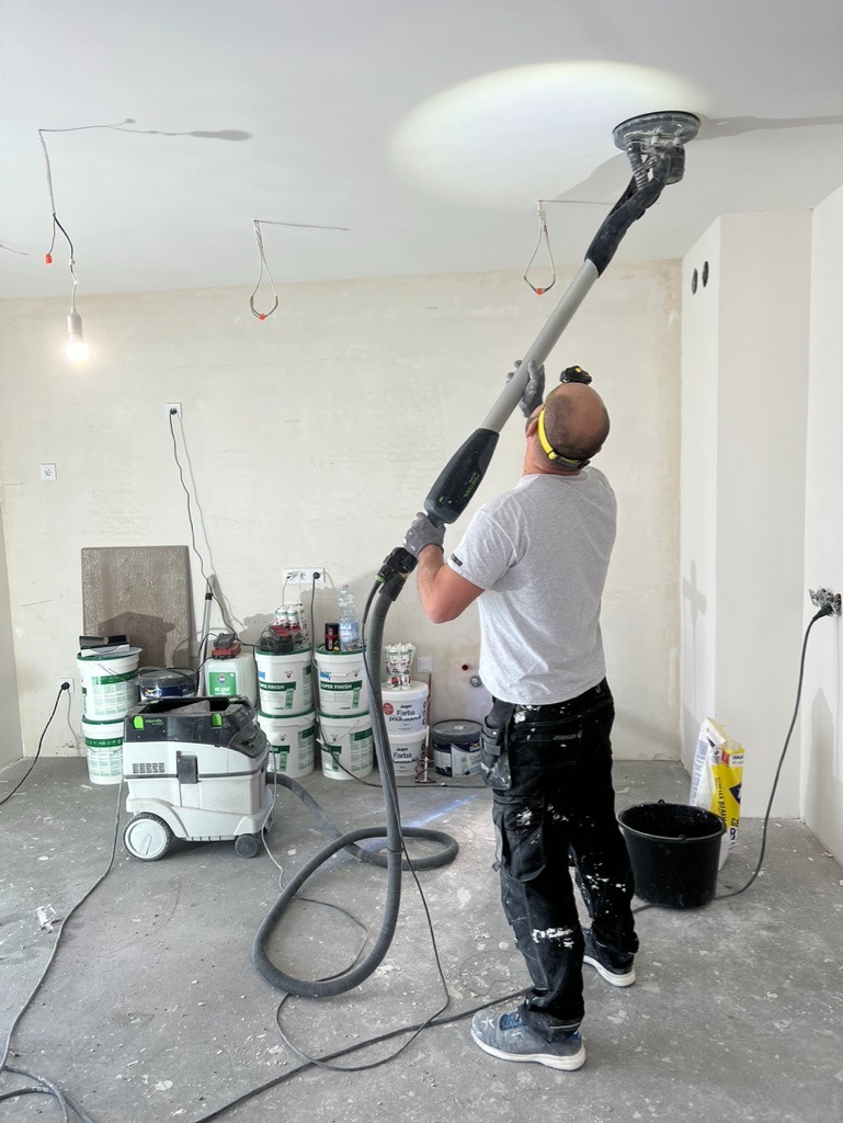 Pracownik szlifuje sufit szlifierką teleskopową w pomieszczeniu w trakcie remontu. Widoczne puszki z farbami, odkurzacz przemysłowy i inne narzędzia.