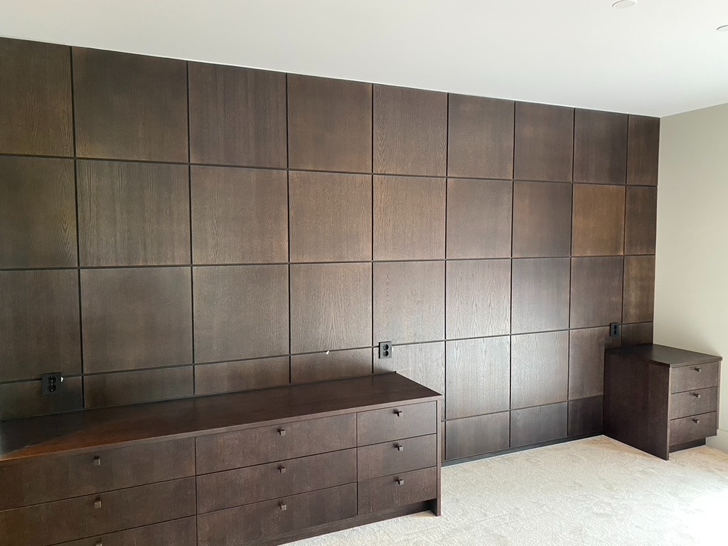 Nowoczesna sypialnia z ciemnobrązową ścianą z paneli drewnianych, dopasowaną komodą i szafkami nocnymi. Minimalistyczny design, jasna podłoga.