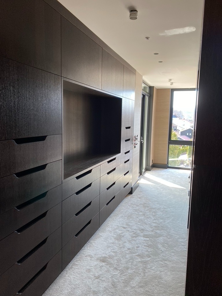 Wnętrze garderoby z ciemnymi, drewnianymi szafami na wymiar, jasną wykładziną i oknem z widokiem na zieleń. Minimalistyczny design, nowoczesne wykończenie.