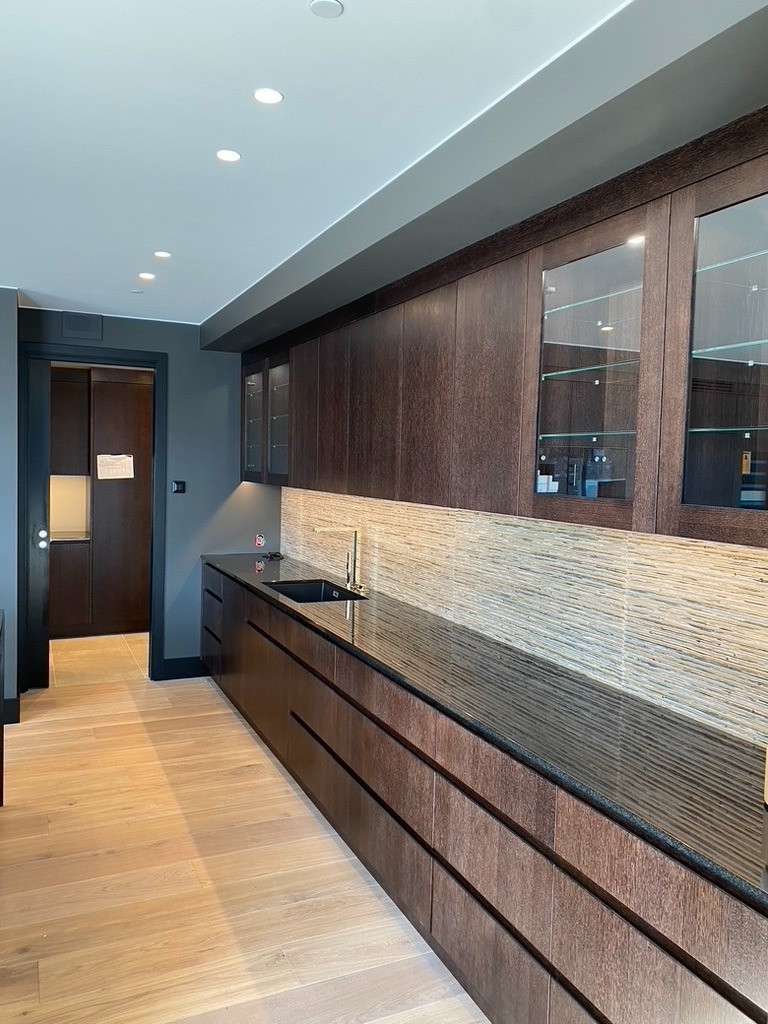 Nowoczesna kuchnia z ciemnymi drewnianymi szafkami, czarnym blatem i jasną podłogą. Uwagę przykuwają szklane witryny górnych szafek i dekoracyjne płytki nad blatem.