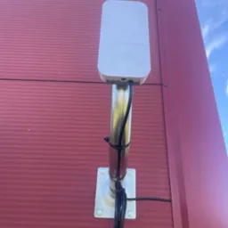 Zewnętrzna antena sieciowa MikroTik zamontowana na metalowej rurze przytwierdzonej do bordowej ściany budynku, kable zabezpieczone opaskami zaciskowymi.