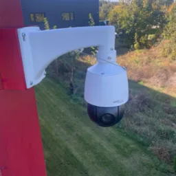 Zewnętrzna kamera monitoringu PTZ zamontowana na czerwonym słupie, z widokiem na zielony trawnik i budynek z ciemną elewacją w tle, w słoneczny dzień.