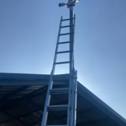 Zewnętrzna instalacja kamer monitoringu na słupie, widoczna drabina oparta o konstrukcję budynku. Błękitne niebo w tle, oświetlenie dzienne.