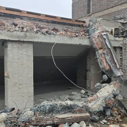 Demolition-repair-company-DCH. - Ciężki sprzęt z hydraulicznym ramieniem kruszy fragment ściany budynku z czerwonej cegły, gruz leży na ziemi, widoczna firma Visser na maszynie.