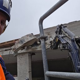 Demolition-repair-company-DCH. - Operator w kasku i kamizelce odblaskowej steruje maszyną do rozbiórki fragmentu betonowego budynku, widoczne zbrojenia i gruz.