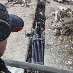 Demolition-repair-company-DCH. - Wnętrze budynku w trakcie prac rozbiórkowych: wąski wykop w podłodze, gruz i resztki materiałów budowlanych, widoczna maszyna do wykuwania bruzd, operator w kabinie.