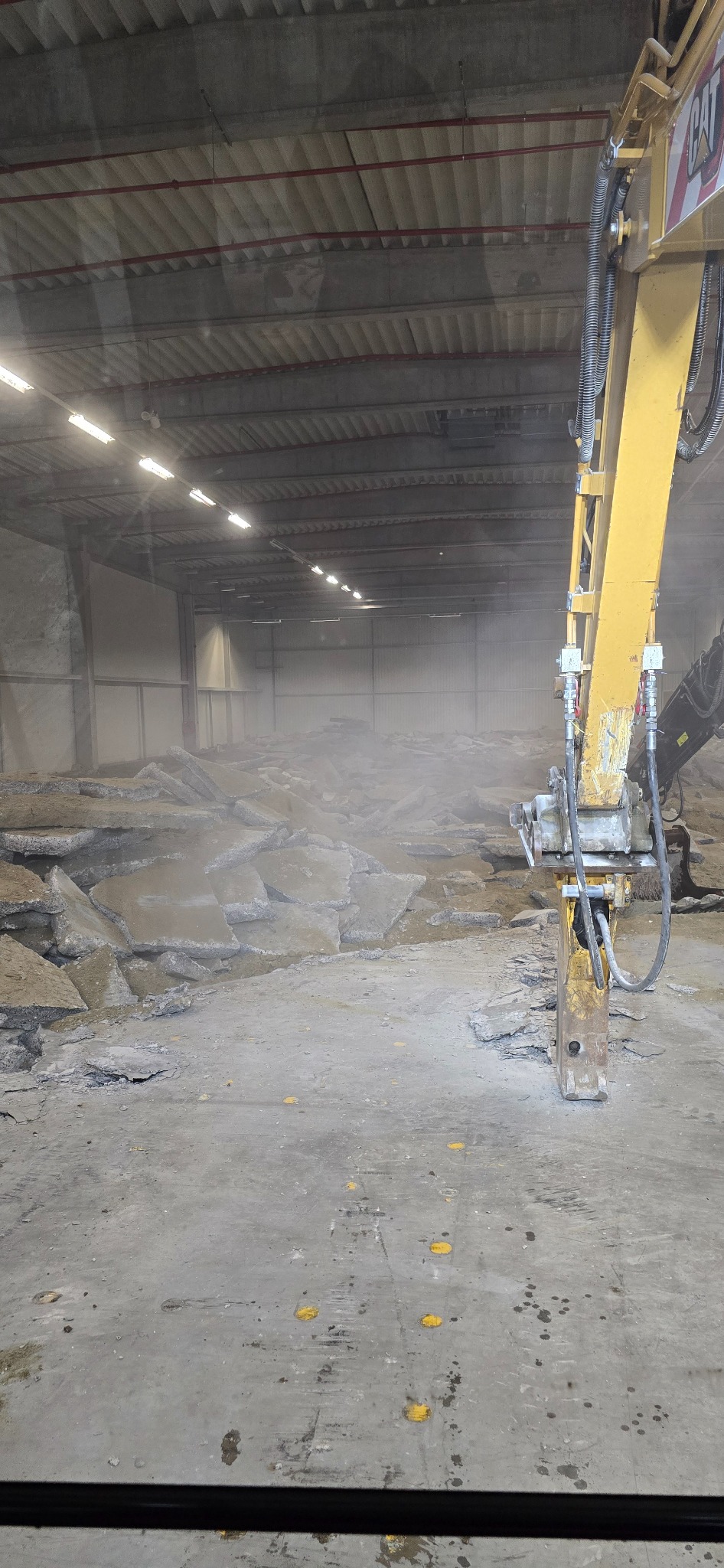 Żółty młot hydrauliczny marki CAT kruszy betonową posadzkę wewnątrz hali przemysłowej, unosząc tumany pyłu.