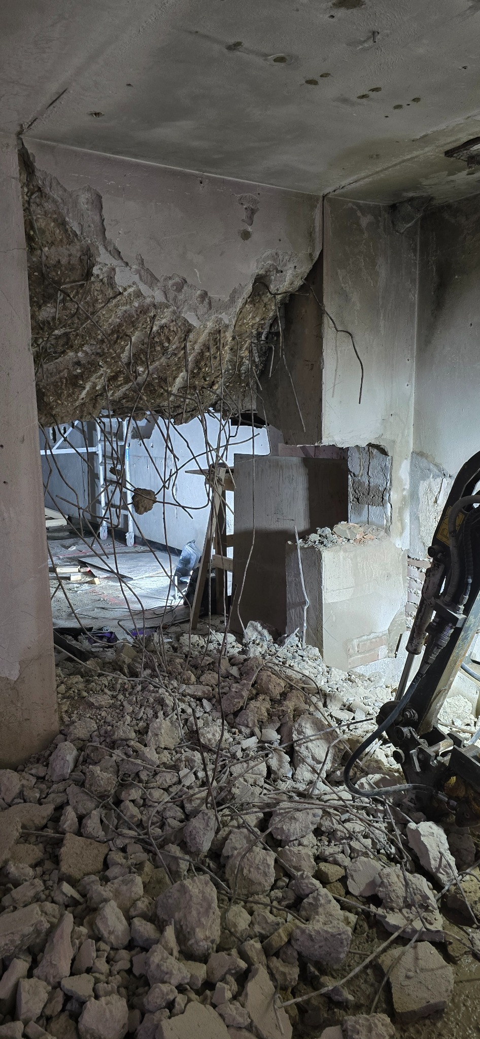 Wnętrze budynku w trakcie rozbiórki, fragmenty gruzu i odsłonięte zbrojenia, w tle drabina i fragment maszyny budowlanej.