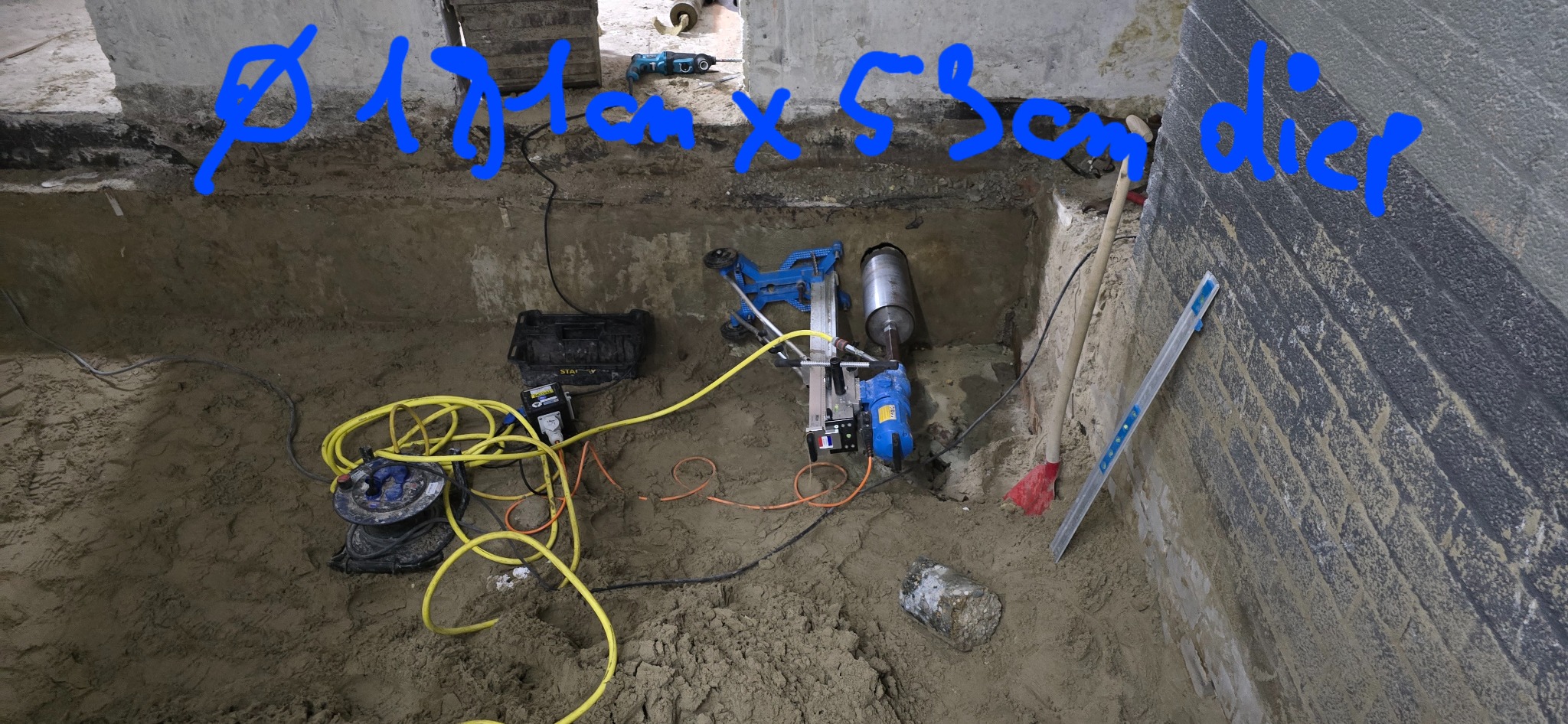 Wykonywanie odwiertu w ścianie fundamentowej za pomocą wiertnicy diamentowej, widoczny wykop, narzędzia i akcesoria.