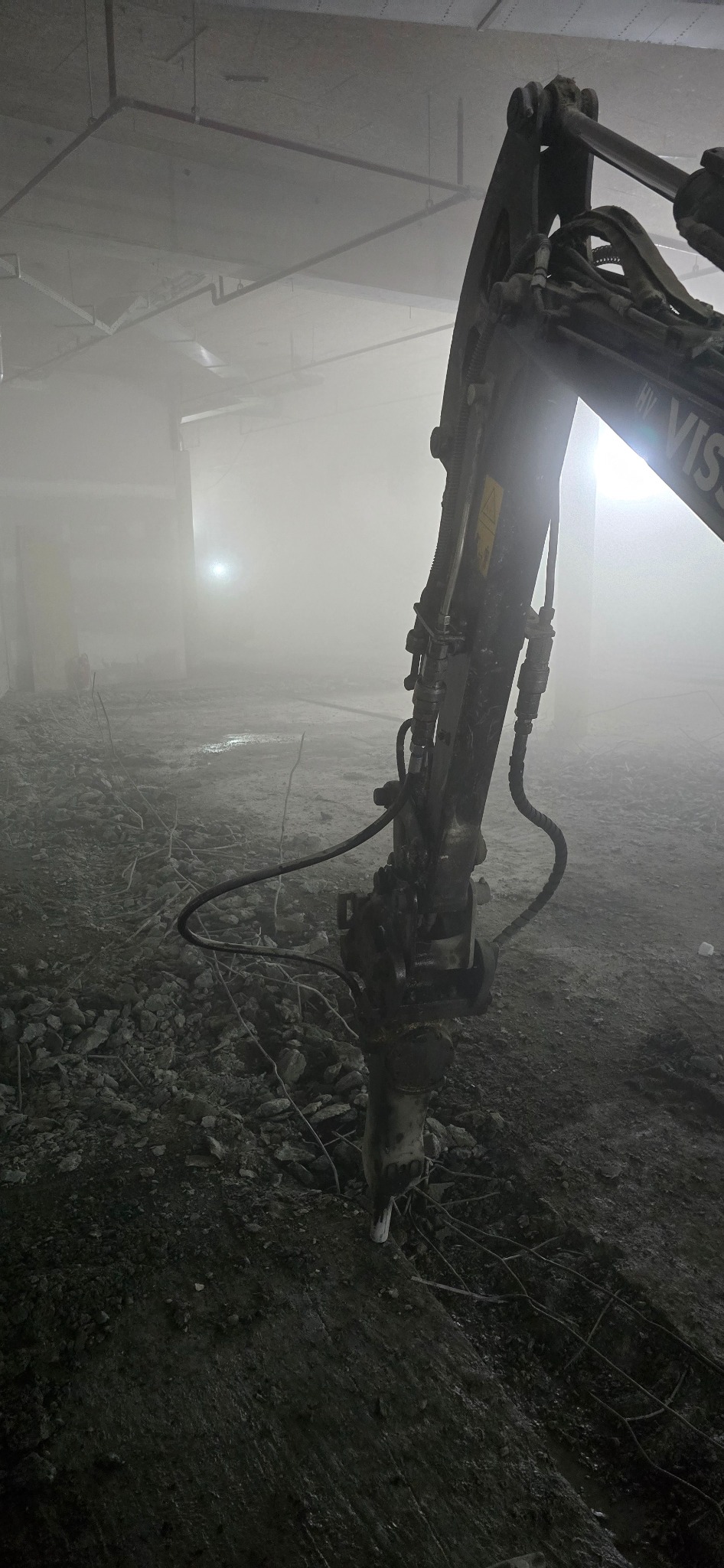 Młot hydrauliczny zamontowany na koparce, kruszący betonową posadzkę w zakurzonym, słabo oświetlonym wnętrzu budynku. Widoczne pozostałości gruzu i wystające pręty zbrojeniowe.