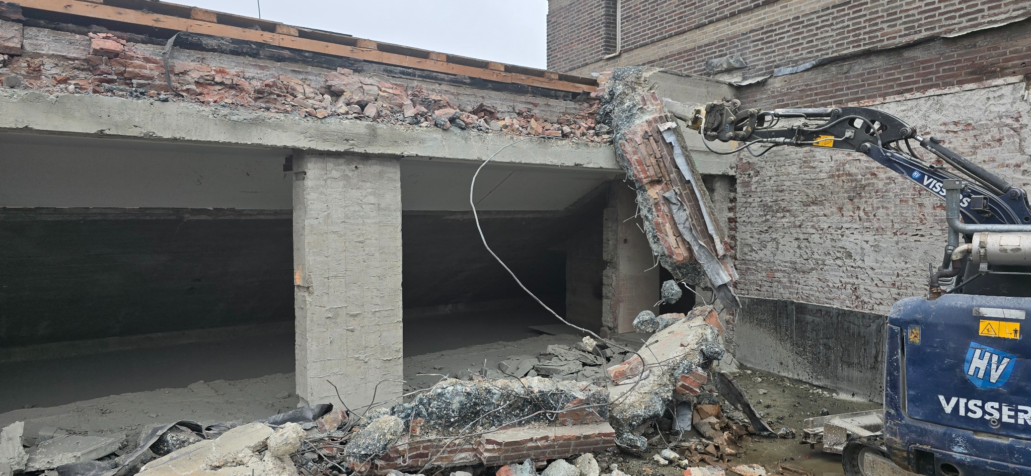 Ciężki sprzęt z hydraulicznym ramieniem kruszy fragment ściany budynku z czerwonej cegły, gruz leży na ziemi, widoczna firma Visser na maszynie.