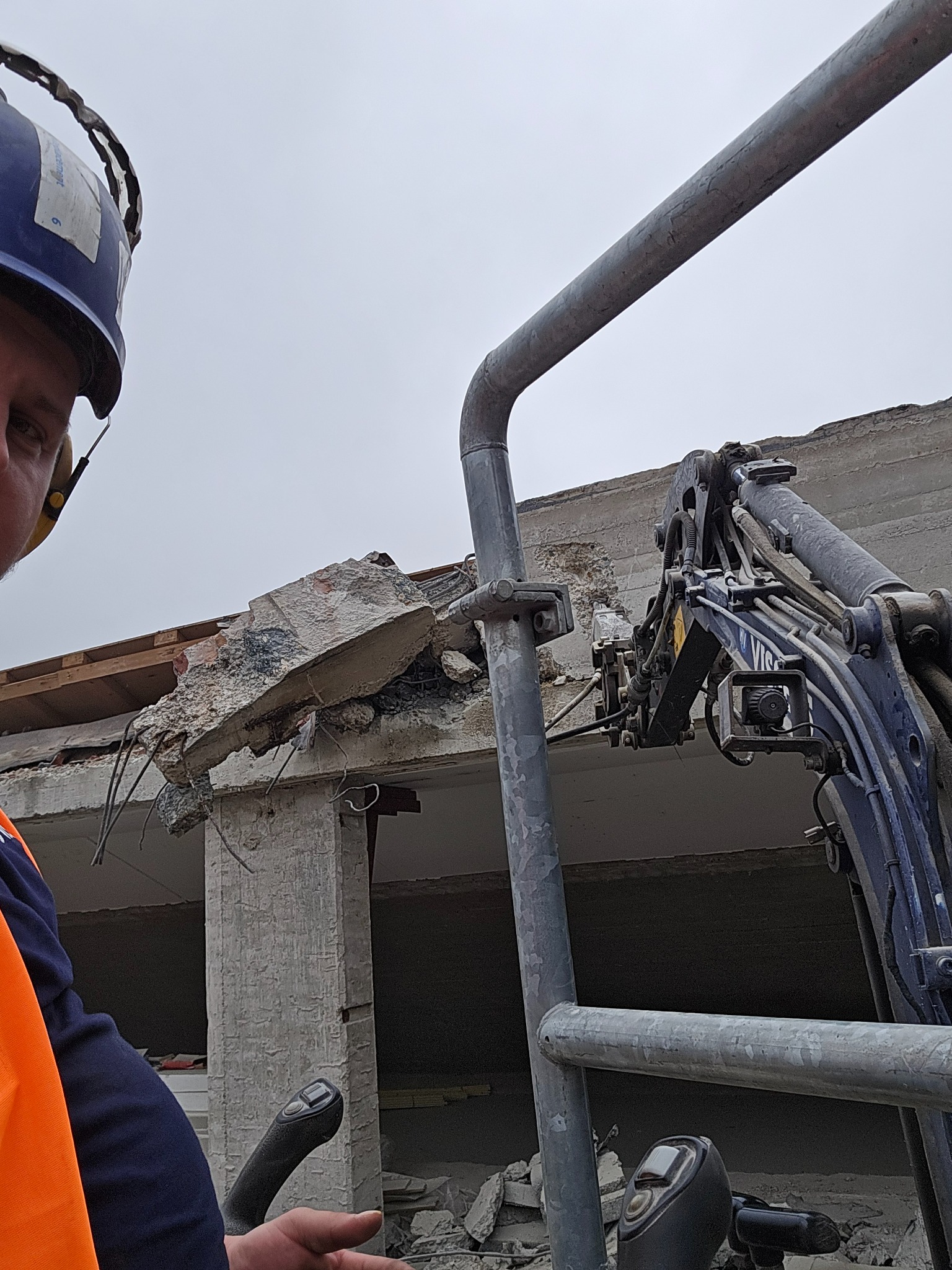 Operator w kasku i kamizelce odblaskowej steruje maszyną do rozbiórki fragmentu betonowego budynku, widoczne zbrojenia i gruz.