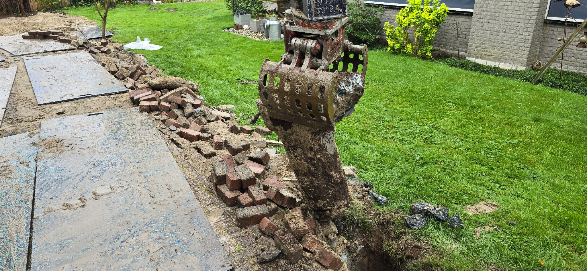 Koparka z ażurową łyżką wydobywa ziemię z wykopu, obok stos cegieł i płyty chodnikowe, trawnik w tle.