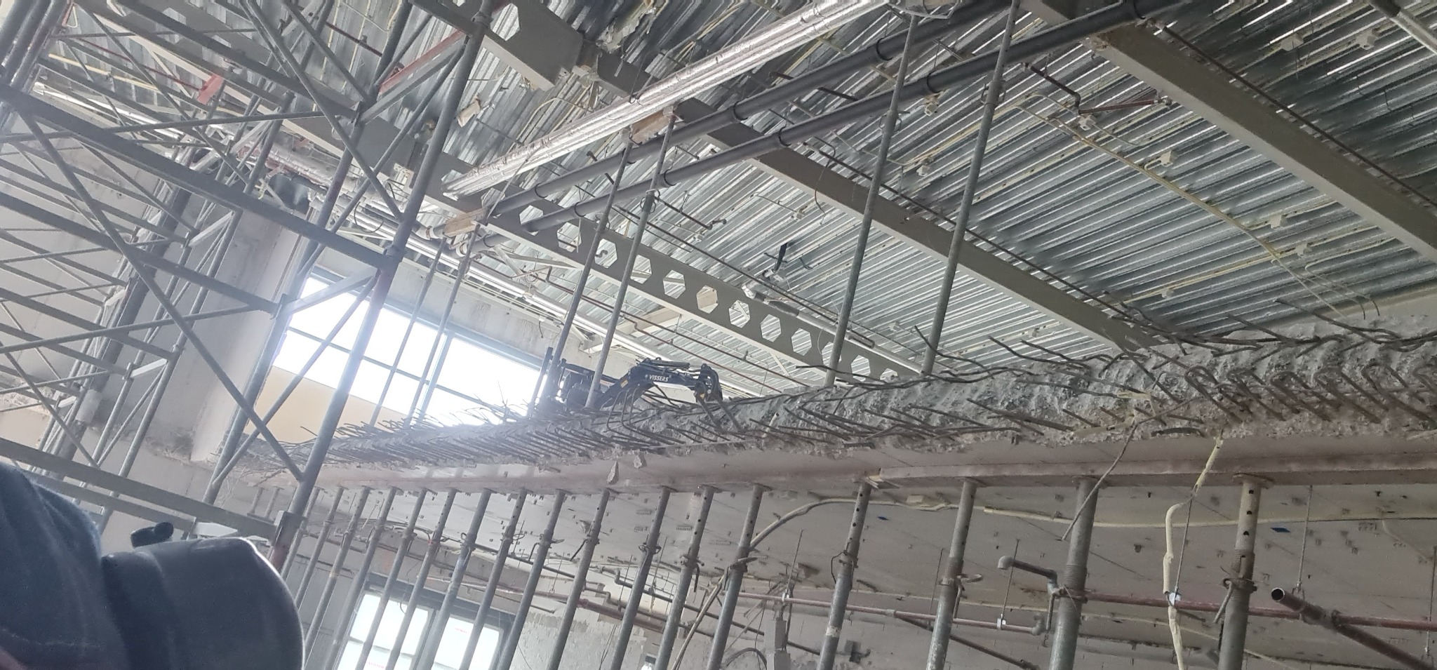 Mini koparka na wysokości, pracująca przy rozbiórce betonowej konstrukcji z wystającymi prętami zbrojeniowymi wewnątrz budynku, widok z dołu na stalowe podpory i metalowy sufit.