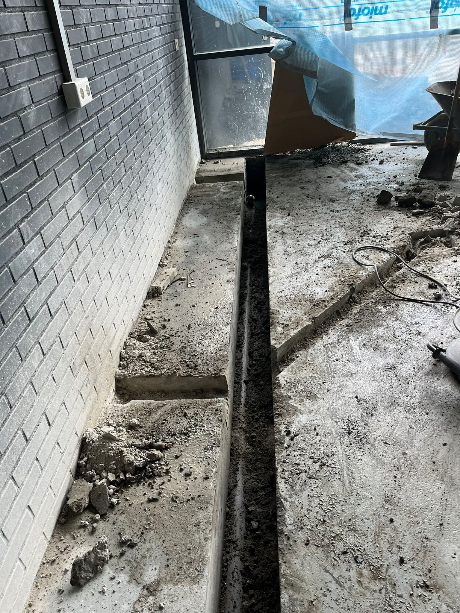 Wąski wykop w betonowej posadzce wzdłuż szarej ściany z cegły klinkierowej, widoczne gruz i narzędzia budowlane w tle.