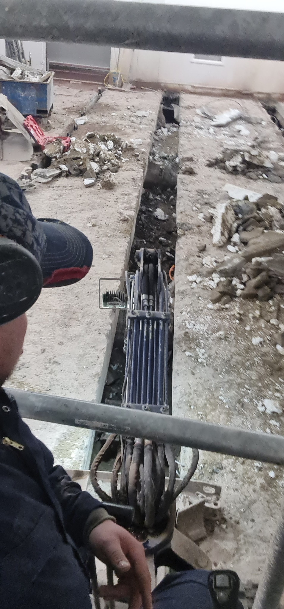 Wnętrze budynku w trakcie prac rozbiórkowych: wąski wykop w podłodze, gruz i resztki materiałów budowlanych, widoczna maszyna do wykuwania bruzd, operator w kabinie.